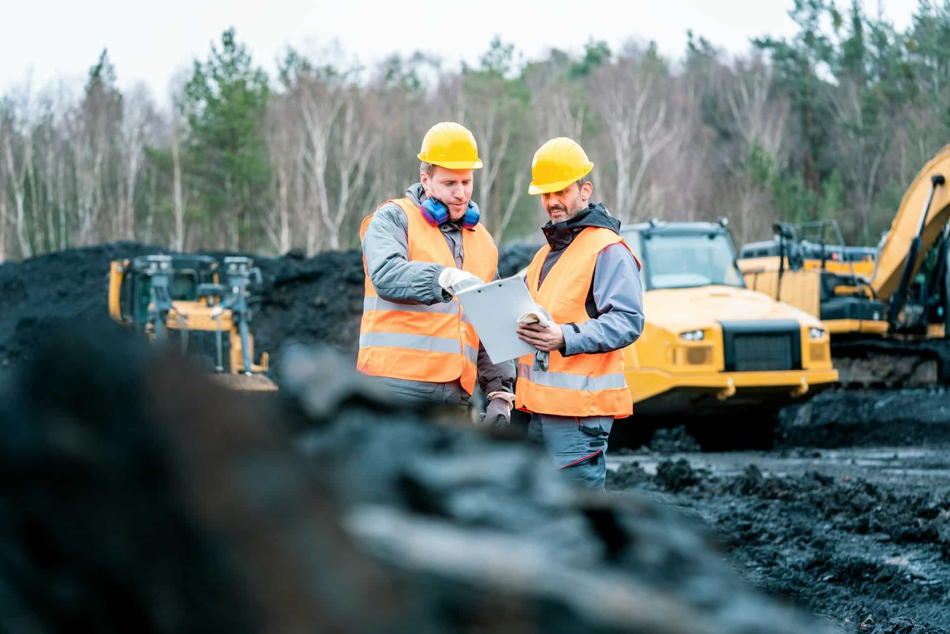 Two construction workers looking at a set of plans outside, in front of a treeline and construction vehicles, representing Certificates of Pending Litigation and land development in BC