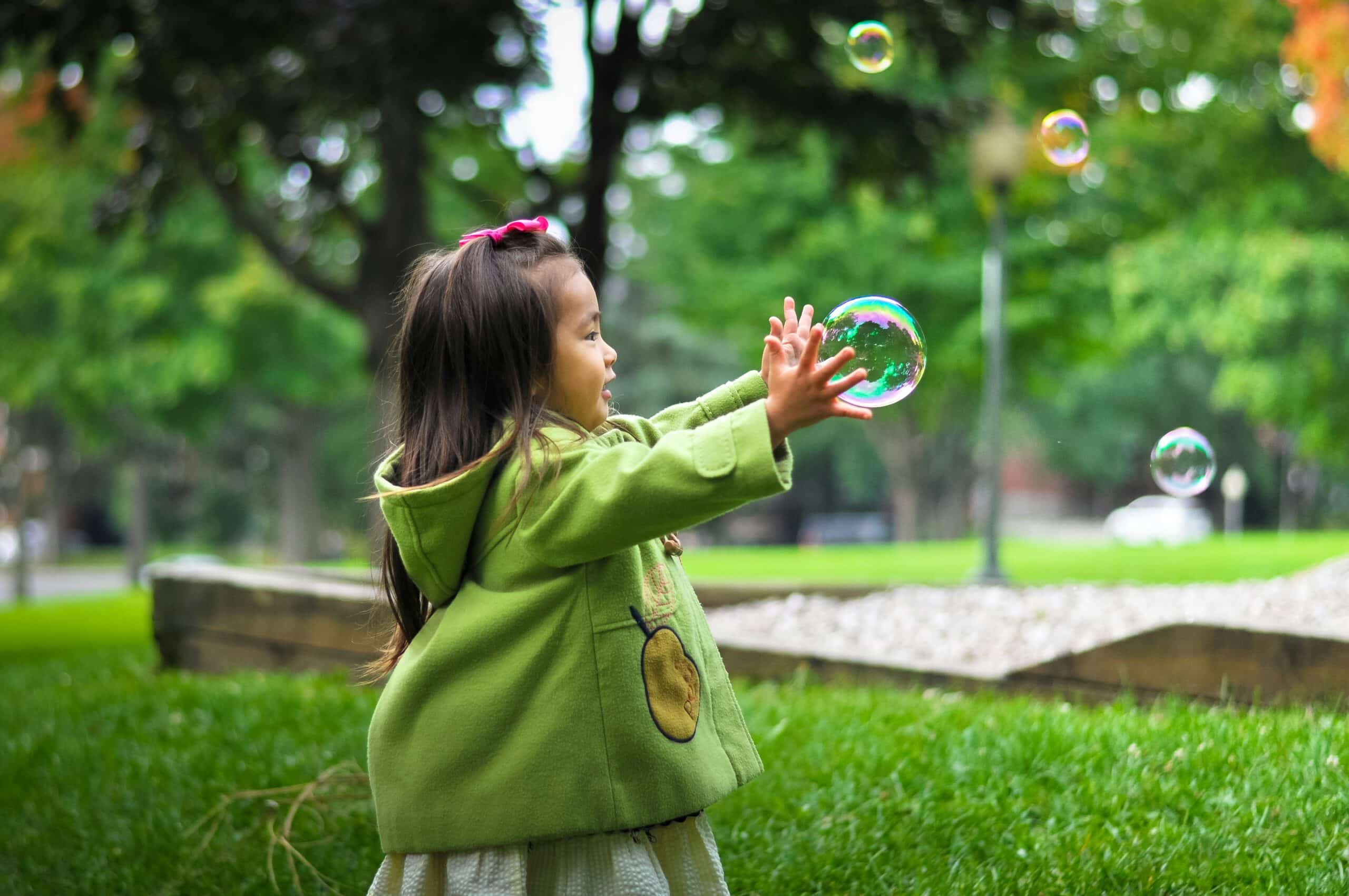 A young girl in a green sweater playing in a lush green park, popping bubbles with her hands, representing Roland Luo and his assistance in high-net-worth family law matters provided in English, Chinese, and French in Vancouver, including matters regarding multi-jurisdictional family property disputes, pension division, family mediation, and child mobility and relocation.